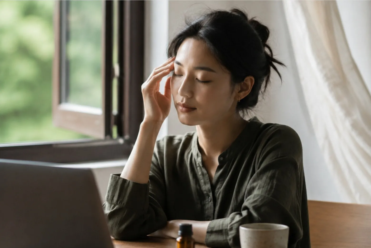 女子在筆電前閉眼，手輕觸太陽穴，桌上有一瓶精油